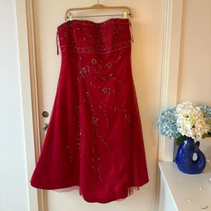Röd axelbandslös balklänning från Jake*s - Snygg röd axelbandslös klänning från Jake*s med glittriga broderade blommor och paljettdetaljer. Klänningen har en vacker A-linjeform och dragkedja i ryggen. Perfekt för bal eller festliga tillfällen. Den är över knäna på mig som är 164cm, och passar perfekt när jag vanligtvis bär strl L
