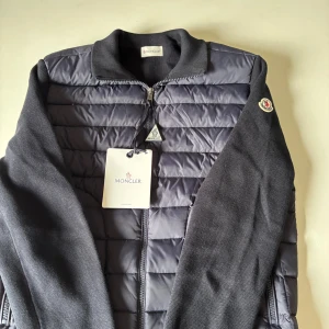 Moncler cardigan - Säljer en svart pufferjacka från Moncler med quiltad front i polyester och stickade ärmar. Jackan har Moncler-logga på ärmen, ribbad krage och dragkedja framtill. Snygg och stilren design som passar perfekt till streetwear och chill vardagsstil. Aldrig andvänd. Sitter lite mer som S men kan även passa M. Priset är ej hugget i sten.