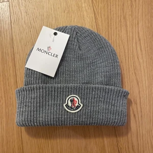 Grå Moncler mössa - Snygg grå mössa från Moncler med ribbad stickning och klassisk Moncler-logga framtill. Perfekt för att hålla värmen och samtidigt se stilren ut. Materialet är mjukt och stickat, vilket gör den skön att bära under kyliga dagar.