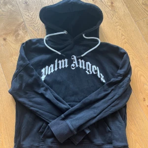 Svart Palm Angels hoodie med tryck - Svart hoodie från Palm Angels med stor vit logotyp på bröstet. Tröjan har huva med vita snören, ribbade muddar och magficka. Materialet är mjuk bomull och polyester, perfekt för chill dagar. Snygg streetwear-vibe och klassisk passform.