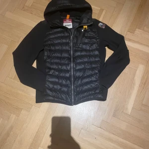 Svart pufferjacka från Parajumpers - Svart pufferjacka från Parajumpers med huva och quiltad front. Jackan har dragkedja, snygga detaljer som orange hängare och patch på ärmen. Materialet är polyester och syntet, perfekt för kyliga dagar. Stilren och sportig look.
