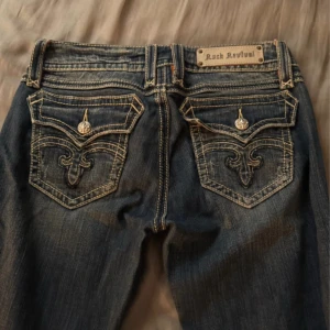 Lågmidjade jeans  - Säljer ett par mörkblå Rock Revival jeans med snygga broderade bakfickor och silverfärgade detaljer. Jeansen har låg midja och raka ben, med slitningar och kontrastsömmar som ger en cool look. Perfekta för dig som gillar statement-jeans! Nypris ca 2000kr 
