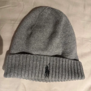 Grå kashmirmössa från Polo Ralph Lauren - Snygg grå mössa från Polo Ralph Lauren i mjuk kashmir. Klassisk ribbad kant med broderad logga framtill. Perfekt för dig som vill ha en stilren och lyxig look under kalla dagar. OBS-Mössan är en barn/bebis mössa 46cm!, läsken är som skala och följer inte med i köpet, kom gärna med prisförslag!