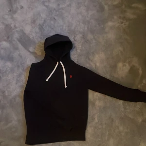 Svart hoodie från Polo Ralph Lauren - Svart hoodie från Polo Ralph Lauren med vit snörning och röd logga på bröstet. Klassisk design med huva och långa ärmar, perfekt för chill dagar. Mjuk bomullskänsla och stilren look som funkar året runt.