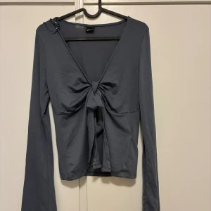 Grå långärmad topp med knyt - Snygg grå långärmad topp med djup v-ringning och knyt framtill. Toppen har en croppad passform och är tillverkad i mjukt material som känns skönt mot huden. Perfekt att styla med jeans eller kjol för en trendig look.