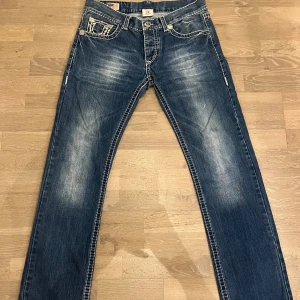 True Religion blå jeans med broderade fickor - Säljer ett par blå jeans från True Religion med snygga kontrastsömmar och broderade bakfickor. Jeansen har klassisk femficksdesign, raka ben och slitningar framtill för en cool look. Perfekt för dig som gillar streetstyle och detaljer. Nypris ungefär 1000kr+ säljer för 200. Byxorna knappt använda så jävligt bra pris