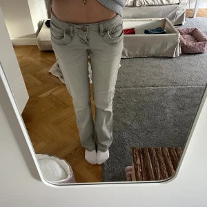 Ljusgrå lågmidjade jeans - Säljer ett par ljusgrå jeans med låg midja och raka ben. Jeansen har klassiska fem fickor, snygga sömdetaljer och en enkel knappstängning framtill. Perfekta för dig som gillar en avslappnad och stilren look.