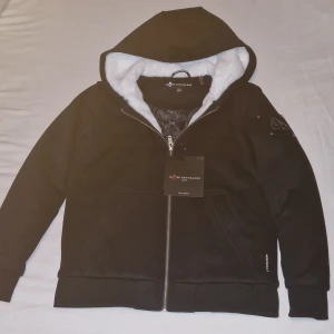Svart tröja Moose Knuckles  barn XL/ XS - Svart tröja från Moose Knuckles i storlek XS med vit fluffig päls på insidan av huvan. Tröjan har dragkedja framtill, två fickor och en snygg metallpatch på ärmen. Perfekt för dig som gillar streetwear och vill ha en varm hoodie med coola detaljer.