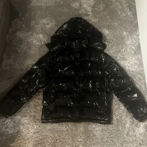 Svart pufferjacka från Trapstar - Säljer en svart, glansig pufferjacka från Trapstar med huva och dragkedja. Jackan har ett stort Trapstar-tryck på ryggen och en rund patch med logga på bröstet. Snygg streetstil och perfekt för kalla dagar. Fylld med syntetmaterial för extra värme.