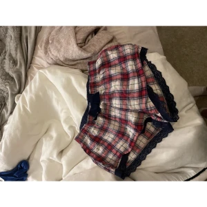 Rutiga shorts med spets och rosett - Säljer ett par rutiga shorts i rött, blått och vitt med mörkblå spetsdetaljer längs kanterna och en söt rosett framtill. Perfekta för chill hemma eller som pyjamasshorts. Mjukt material och bekväm passform.