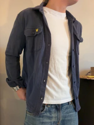 Mörkblå overshirt från Lyle & Scott - Säljer en mörkblå overshirt från Lyle & Scott Junior med klassisk krage, två bröstfickor med knapp och den ikoniska gula logon på vänster ficka. Skjortan har långa ärmar och knäppning framtill. Perfekt att slänga på över en t-shirt för en chill look.