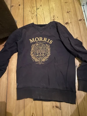 Svart sweatshirt från Morris R Club - Svart sweatshirt från Morris R Club med stort guldfärgat tryck på bröstet med universitetsinspirerat emblem. Tröjan har rund hals, ribbade muddar och är långärmad. Perfekt för dig som gillar klassisk och sportig stil.
