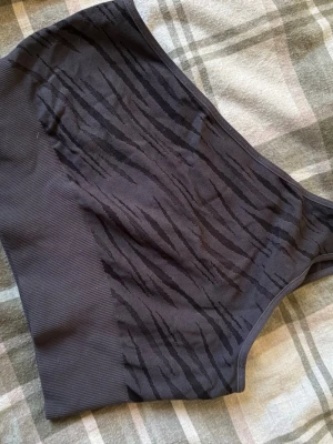 Mörkgrå zebra bandeau topp - Säljer en mörkgrå bandeau topp från Soccer med zebramönster. Toppen är ärmlös och har en bred ribbad kant nedtill för extra komfort. Materialet är 96% polyester och 4% elastan, vilket gör den stretchig och skön. Perfekt för träning eller chill.