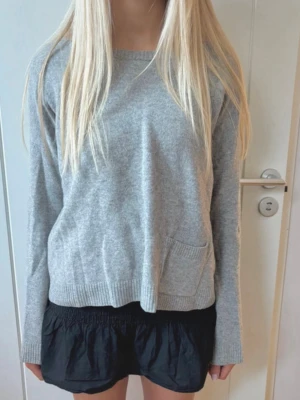 Grå stickad ulltröja - Mysig grå stickad tröja med rund hals och en liten ficka framtill. Tröjan har långa ärmar och ribbade muddar vid hals, ärmslut och nederkant. Vet ej storlek men passar S. Materialet är ull och kaschmir. Märket är Marc O’Polo.