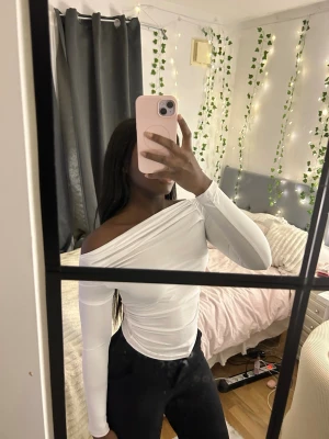 Vit offshoulder topp - Säljer en vit, tight offshoulder topp med lång ärm. Toppen har en snygg omlott-detalj över axeln och är figurnära med stretchigt material. Perfekt för dig som gillar stilrena och trendiga plagg med en modern twist.