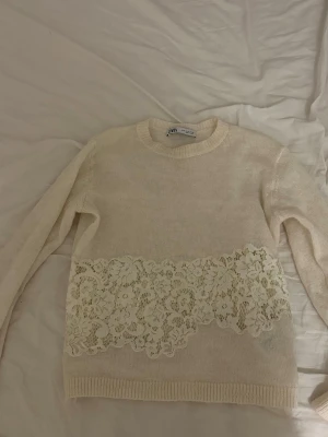 Beige stickad tröja med spets från Zara - Säljer en beige stickad tröja från Zara med lång ärm och rund hals. Tröjan har en bred dekorativ spetsdetalj med blommigt mönster över magen, vilket ger en unik och feminin touch. Perfekt för dig som gillar mjuka färger och snygga detaljer.