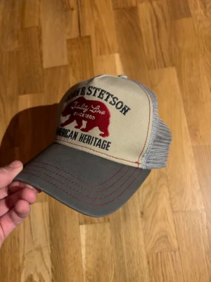 Grå och beige Stetson keps  - Säljer en snygg truckerkeps från Stetson med beige front, grå skärm och mesh på sidorna. Broderad röd björn och texten 'American Heritage' framtill. Justerbar snapback baktill för perfekt passform. Perfekt för dig som gillar klassisk amerikansk stil.