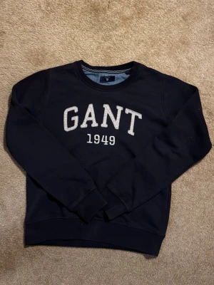 Mörkblå sweatshirt från GANT - Mörkblå sweatshirt från GANT med stort broderat vitt tryck 'GANT 1949' på bröstet. Tröjan har rund hals med ribbad kant och långa ärmar.