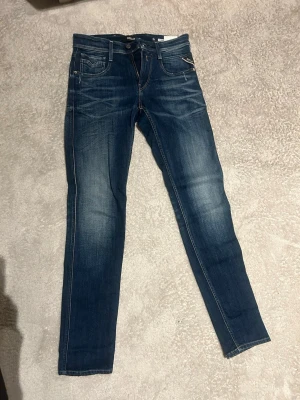Replay Anbass jeans blå denim - Säljer ett par snygga Replay Anbass jeans i klassisk blå denim med lätt slitning och kontrastsömmar. Modellen har fem fickor, dragkedja och knapp framtill samt Replay-logga på fickan. Passformen är slim och materialet är stretchigt jeans.