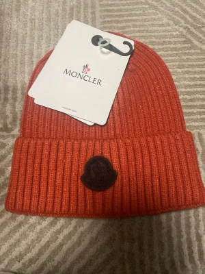 Ingen väst på bilderna - Bilderna visar en orange stickad mössa från Moncler, inte en väst. Mössan har ribbad struktur och ett Moncler-märke framtill.