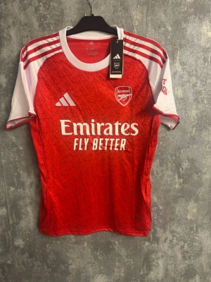 Arsenal matchtröja storlek M - Snygg Arsenal matchtröja från Adidas i rött och vitt med vita ränder på axlarna och klubbmärke på bröstet. Tröjan har ett subtilt mönster över hela framsidan och logga från Emirates. Perfekt för dig som älskar fotboll och vill visa ditt stöd för Arsenal.