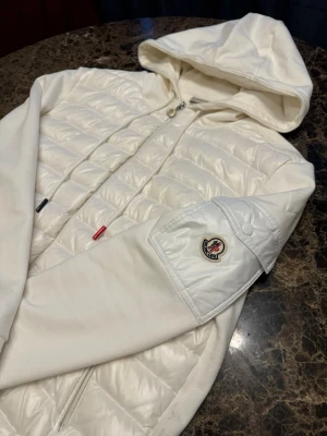 Vit pufferjacka från Moncler med huva - Snygg vit pufferjacka från Moncler med quiltad framsida och huva. Jackan har dragkedja, snörning i halsen och en ficka med Moncler-logga på ärmen. Materialet är polyester och bomull, med en clean och modern look.