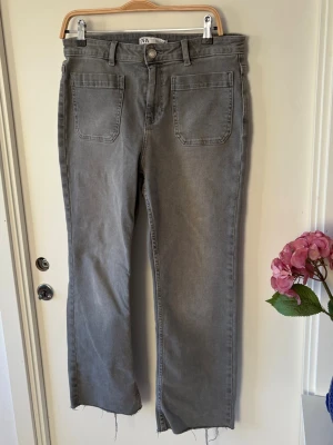 Grå raka jeans från Zara - Säljer ett par grå jeans från Zara i storlek 40. Modellen har raka ben, mid waist och råa, fransiga benslut. Jeansen har stora fickor både fram och bak samt klassisk knapp och dragkedja. Materialet är jeans och färgen är mörkgrå.