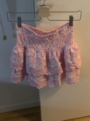 Rosa volangkjol från Kappahl - Supersöt kort kjol från Kappahl i vitt och rosa med broderade detaljer och volanger. Kjolen har ett elastiskt midjeband och ett unikt mönster som ger den en lekfull vibe. Perfekt för dig som gillar färg och snygga detaljer. perfekt för utomlands eller nu till sommaren