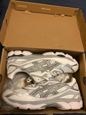 Vita och grå Asics sneakers - Säljer ett par snygga Asics sneakers i vitt och grått med coola detaljer och meshpaneler. Skorna har snörning och en platt sula som ger skön komfort. Perfekta för dig som gillar sportig stil och vill ha något som funkar till många outfits.