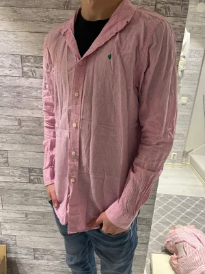 Rosa linneskjorta från Morris - Säljer en rosa skjorta från Morris i ren linne, med klassisk krage och knappar framtill. Skjortan har lång ärm och en liten grön broderad detalj på bröstet. Perfekt för dig som gillar en chill och stilren look.