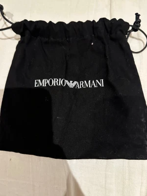 Svart tygpåse från Emporio Armani - Säljer en svart tygpåse från Emporio Armani med vit logotyp framtill. Påsen har snörning upptill och är perfekt för att förvara mindre accessoarer eller smycken. Enkel och stilren design som passar dig som gillar klassiska märken.
