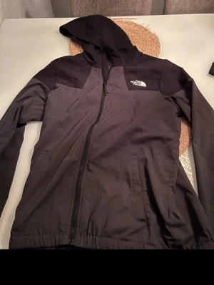 Svart vindjacka från The North Face - Säljer en svart vindjacka från The North Face med huva och dragkedja framtill. Jackan har två sidofickor och en stilren logga på bröstet. Perfekt för dig som vill ha en lätt och smidig jacka till vardags. Materialet är syntet och jackan har en normal passform.
