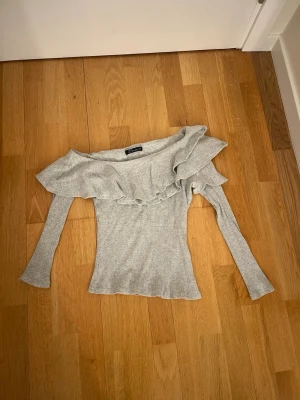 Grå offshoulder topp med volanger - Säljer en ljusgrå ribbad topp från Simplee i storlek S. Modellen är offshoulder med lång ärm och har en bred volang längs halsringningen. 