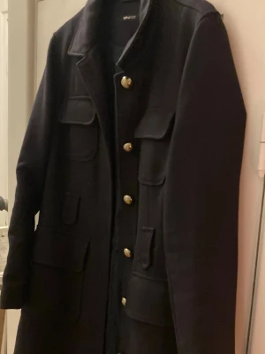 Svart kappa med guldknappar från Ginatricot - Snygg svart kappa från Ginatricot med flera fickor och guldiga knappar framtill. Klassisk krage och långärmad modell, perfekt för dig som gillar stilrena detaljer. Materialet känns mjukt och har en ullig känsla. Passar dig som vill ha en cool och tidlös look.