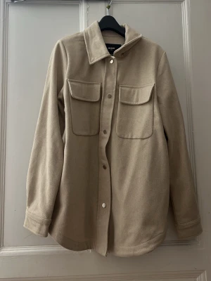 Beige overshirt från Bik Bok - Säljer en beige overshirt från Bik Bok med klassisk krage, två stora bröstfickor och tryckknappar framtill. Jackan har lång ärm och är gjord i mjukt ullmaterial som ger en chill och clean look. Perfekt att slänga över en hoodie eller t-shirt.