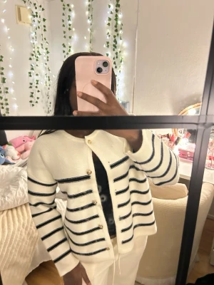 Vit och marinblå randig kofta - Säljer en snygg vit kofta med marinblå horisontella ränder och guldfärgade knappar. Koftan har rund halsringning och långa ärmar. Perfekt att slänga på sig över en topp för en chill och stilren look. 