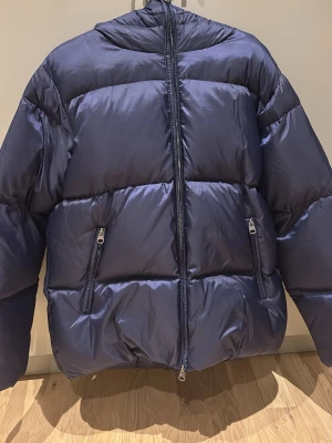 Blå dunjacka från Stories S - Snygg mörkblå dunjacka från Stories i storlek S. Jackan är lite oversized, huva, två fickor med dragkedja och en stor dragkedja framtill. Materialet är dun och polyester, perfekt för kalla dagar. 