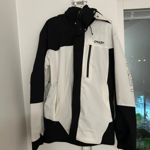 Svartvit fodrad skidjacka från Oakley - Oakley fodrad skidjacka i svart och vitt med coolt blockmönster och logga på bröstet och ärmen. Jackan har huva, snörning och dragkedja framtill samt en bröstficka med zip. Perfekt för dig som vill se cool ut i skidbacken.