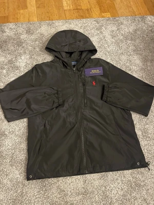 Svart vindjacka från Polo Ralph Lauren - Svart vindjacka från Polo Ralph Lauren med huva, dragkedja och elastiska muddar. Jackan har en röd broderad logga på bröstet och en bröstficka med dragkedja. Tillverkad i glansig polyester för en clean och sportig look. Byten funkar