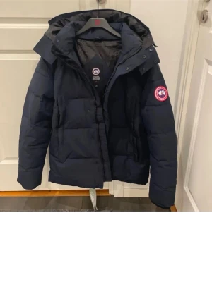 Mörkblå pufferjacka från Canada Goose - Säljer en mörkblå pufferjacka från Canada Goose med huva och klassisk logopatch på ärmen. Jackan har dragkedja och knappar framtill, samt stora fickor. Perfekt för kalla dagar och har en schysst, lite oversized look.