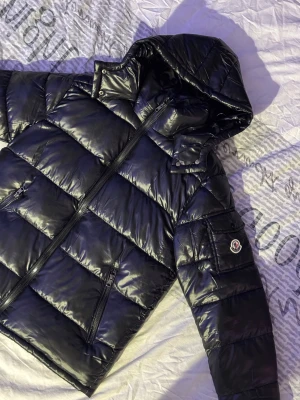 Svart Moncler pufferjacka med huva - Säljer en svart pufferjacka från Moncler med glansig finish och huva. Jackan har dragkedja framtill, två sidofickor med dragkedja och en ficka med Moncler-logga på ärmen. Insidan har ett coolt serietidningsmotiv. Perfekt för kalla dagar!