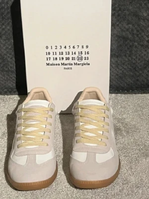 Maison Margiela  - Maison Margiela sneakers i beige och vit med ljusgula snören och gummisula. Skorna har en klassisk rund tå och platt sula, med paneler i mocka och skinn. Snygg retrostil som funkar till många outfits. Box medföljer. Storleken är 43 men passar best för 42