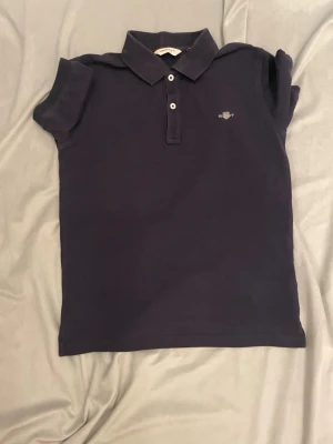 Mörkblå pikétröja från GANT - Snygg mörkblå pikétröja från GANT med klassisk krage och två knappar framtill. Tröjan har korta ärmar och ett litet broderat GANT-logo på bröstet. Materialet är mjuk bomull som känns skönt mot huden. Perfekt för en chill och stilren look. Den har blivit ganska blek på grund av styckning jag har strykt den ganska mycke men syns inte så mycket när man har på sig den. Storlek 152!