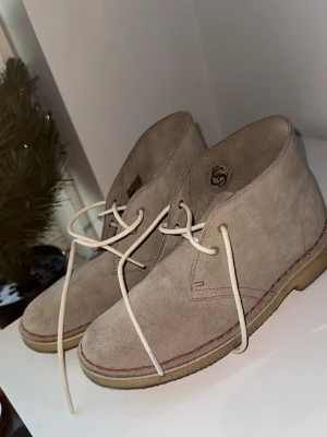 Beige mocka Clarks ökenkängor - Säljer ett par klassiska Clarks ökenkängor i beige mocka med vita snören och kontrastsömmar. Skorna har rund tå, platt sula och är tillverkade i mjukt mockamaterial. Perfekta för dig som gillar stilrena och tidlösa boots. Storlek 37/38. 