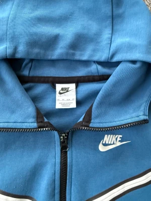 Blå Nike tech - Nike vindjacka i blått med vit logga på bröstet och vita detaljer över bröstet. Jackan har huva, hel dragkedja och fickor fram samt en extra ficka med dragkedja på ärmen. Tillverkad i mjuk polyester, perfekt för chill och streetwear. Liten ytlig reva på högra armen, se bild. I övrigt mycket bra skick.