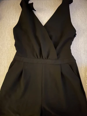 ZARA Svart byxdress med spets och omlott - Säljer en svart byxdress med omlott framtill och breda axelband som knyts. Baksidan har snygga spetsdetaljer som ger en extra touch. Materialet är mjukt och har en lätt stretch.