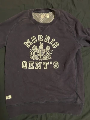 Mörkblå sweatshirt från Morris Gents - Snygg mörkblå sweatshirt från Morris Gents med stort tryck och emblem på bröstet. Tröjan har rund hals, ribbade muddar och är tillverkad i mjuk bomull. Perfekt för dig som gillar klassisk stil med en modern twist.
