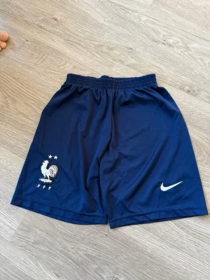Blå Nike shorts Frankrike FFF 164 - Säljer ett par marinblå Nike shorts med Frankrikes landslagslogga (FFF) och två stjärnor. Shortsen har elastisk midja och är tillverkade i lätt polyester, perfekta för fotboll eller träning. Snygg och sportig design med vit Nike-logga på ena benet.