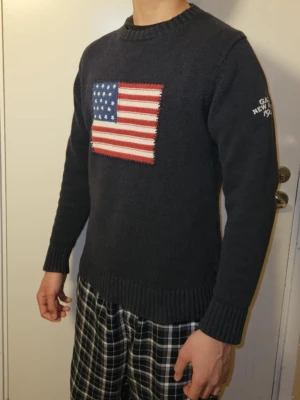 Mörkblå stickad tröja från GANT - Säljer en mörkblå stickad tröja från GANT med amerikansk flagga på bröstet och broderad text 'GANT NEW HAVEN 1949' på ärmen. Tröjan har rund hals, ribbade muddar och lång ärm. Perfekt för dig som gillar klassisk college-stil.