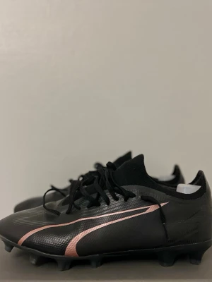 Svarta fotbollsskor från Puma - Säljer ett par svarta fotbollsskor från Puma med snygga roséguld detaljer på sidan. Skorna har snörning och en strumpliknande krage för extra komfort. Yttermaterialet är syntet och sulan har fasta dobbar som ger bra grepp på planen.